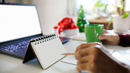 close up shot, hand holding pencil taking note on blank calendar working with laptop, red hat, christmas tree for decoration on desk, christmas holidays concept