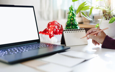 close up shot, hand holding pencil taking note on blank calendar working with laptop, red hat, christmas tree for decoration on desk, christmas holidays concept