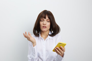 A thoughtful young woman in a white shirt checks a bright yellow smartphone, standing against a plain background as she explores apps, messages, or ideas with calm focus and poised curiosity.