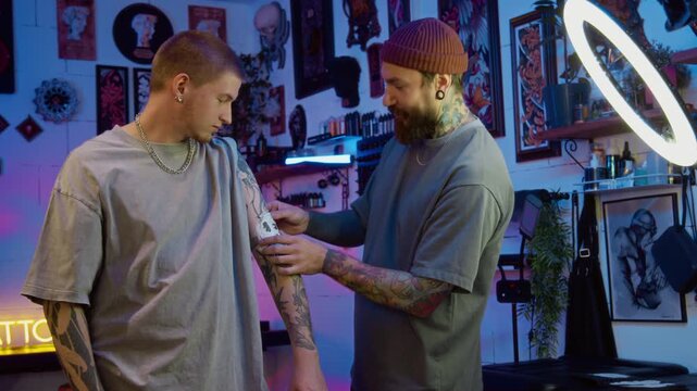 Handheld medium shot of professional bearded young Caucasian male tattooist applying sketch on clients arm, discussing details in cozy studio decorated with posters and ambient lighting
