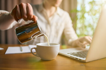 Man pouring milk into coffee cup while working on laptop. Concept of workplace routine, productivity visuals, remote work materials, and subtle beverage-focused content for lifestyle and office themes