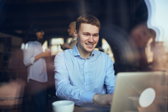 Man typing on laptop in office with reflections and colleagues talking behind. Concept of hybrid work rhythm, deep-focus tasks, urban productivity, and balanced social surroundings.