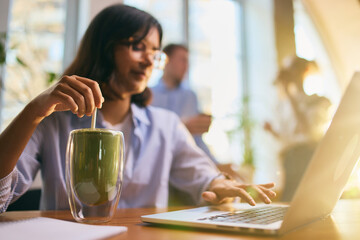 Young woman working on laptop while stirring matcha in bright workspace. Concept of balanced digital routines, healthy drink habits, energizing workflow moments, and wellbeing-focused office visuals.