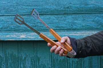 A hand holds one large metal kitchen tongs with a brown wooden handle against a green wall on a summer street