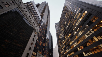 Tall buildings stretch upward in a vibrant cityscape as warm lights begin to glow at twilight. The urban setting exudes a sense of energy and life amidst towering architecture.