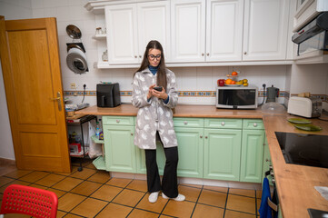 Teenage girl wearing glasses using smartphone while standing in a cozy, well-organized kitchen at home