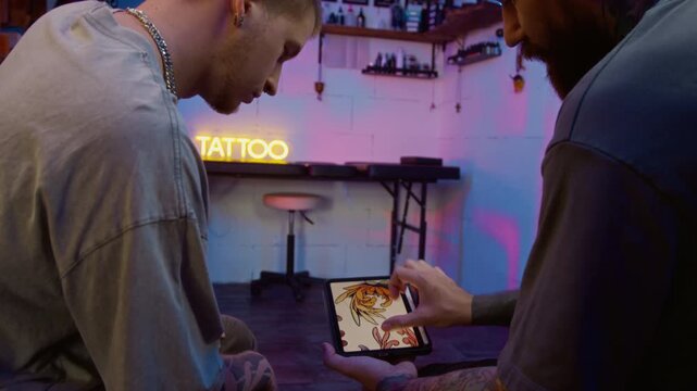 Handheld shot of bearded young Caucasian male tattooist talking to young male client choosing his colored tattoo design on tablet screen in modern studio with ambient neon lighting