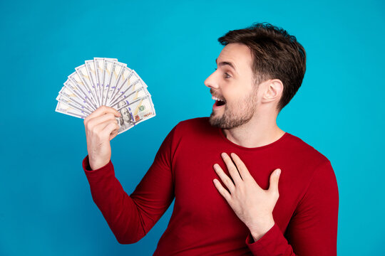 Man in red sweater smiles as he fans a stack of cash against a blue background for a energetic and hopeful lifestyle and shopping themed image