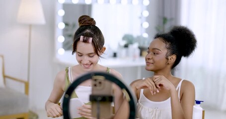 asian teenage girl holding mirror while african american teenage girl talking during beauty tutorial video shoot at home showing skincare content influencer routine with fun and friendship lifestyle