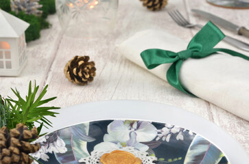 Christmas decoration on wooden table, preparations for dinner, table setting with green bow, cutlery, plates and glass, small white house, lights, moss, cones and evergreen branches.