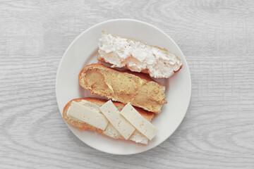 Three sandwiches with pâté, butter, and cheese on a white plate. White wooden background, top view.