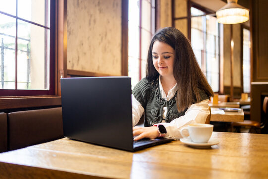 A young girl with Down syndrome is working in a modern cafe using a laptop computer.The girl is writing on the laptop.Concept of trisomy 21. - Powered by Adobe