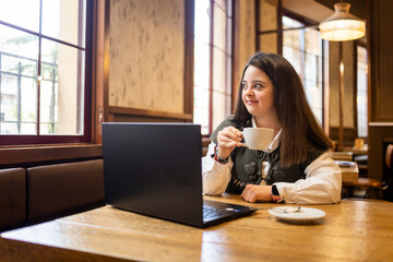 A young girl with Down syndrome is working in a modern cafe using a laptop computer.The girl looking out the window while holding a cup of coffee.Concept of trisomy 21.
