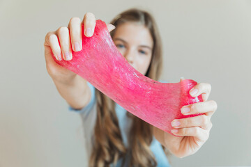 Close-up of hands stretching bright pink slime towards the camera, with a smiling girl in the background. Sensory toy