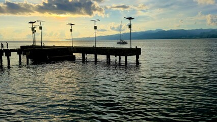 Obraz premium Serene wooden pier extends into calm waters with distant mountains and a boat under a cloudy sky at sunset