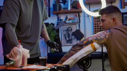 Side view shot of bearded young Caucasian male tattoo artist applying after care products on fresh tattoo sleeve of his young male client, using specialized cleansers to promote fast healing