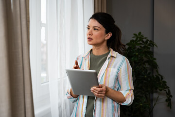 Fototapeta premium Focused woman standing by window review important digital notes on her tablet organizing task planning personal goals learning new skills managing her everyday responsibilities with calm concentration