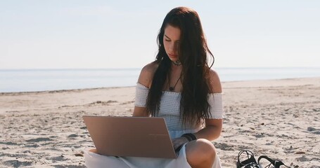 Young woman in summer dress sits on sandy beach with laptop, looks at screen and typing, chatting, engage in freelance work or studying process on nature. Telework, modern tech use, blogging concept