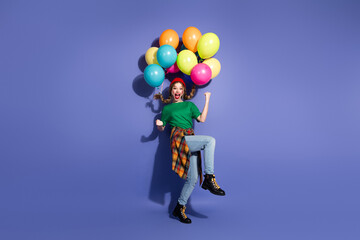 Joyful young girl with colorful balloons leaps against purple background in casual street wear