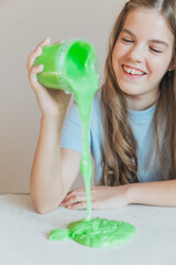 Smiling girl pouring vibrant green slime from a container onto the table. Trendy DIY and fun sensory play activity
