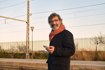Mature adult man smiling looking at camera using smartphone at train station