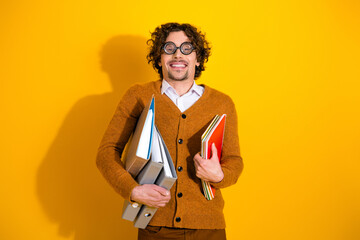 Young man with glasses in a brown cardigan holding notebooks and folders against a bright yellow background for education and lifestyle advertising