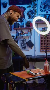 Vertical handheld shot of young Caucasian male tattoo artist in black latex gloves pouring ink in separate ink caps on table, preparing pigment for tattooing session in modern studio