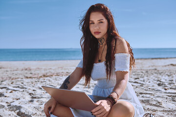 Young 20s beautiful freelancer woman in white summer dress sitting on sandy beach with laptop, posing, looking at camera. Teleworking, online studying, blogging concept