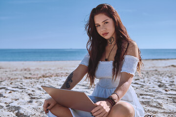 Young 20s beautiful freelancer woman in white summer dress sitting on sandy beach with laptop, posing, looking at camera. Teleworking, online studying, blogging concept