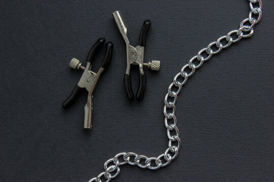 Metal nipple clamps with a silver chain arranged on a dark surface. Clean studio product shot for adult accessories