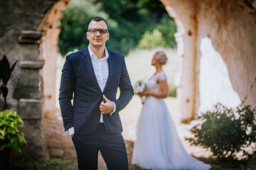 Stylish man in a formal suit stands confidently in a picturesque outdoor setting, while a bride in a flowing gown holds a bouquet, capturing a romantic wedding moment