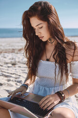 Young woman in summer dress sits on sandy beach with laptop, looks at screen and typing, chatting, engage in freelance work or studying process on nature. Telework, modern tech use, blogging concept