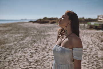 Young serene woman in white dress enjoy warmth and sunny weather with eyes closed stands on sandy beach, feel free and happy. Meditative moment, sensing unity between body, mind, and the earth concept