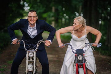 Couple in formal attire racing on vintage bicycles through a lush green forest path, showcasing playful competition and joyful spirit in a unique outdoor setting