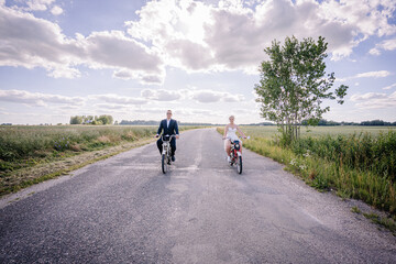 Obraz premium Couple riding bicycles on a scenic country road, surrounded by lush greenery and open fields, under a bright blue sky with fluffy clouds, capturing a joyful moment of togetherness