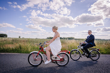 Obraz premium Bride in white dress riding a bicycle alongside groom in suit on a sunny day, surrounded by lush green fields and blue sky, capturing a joyful moment of celebration