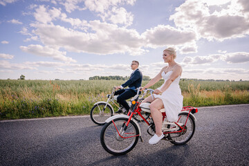 Fototapeta premium Couple riding bicycles on a scenic road, dressed in formal and casual attire, surrounded by lush green fields and a bright blue sky, capturing a joyful moment of togetherness