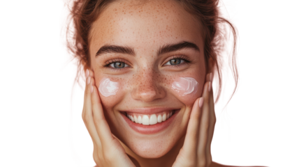 Young woman with freckles smiles as she applies cream to her face, enjoying her skincare routine in a bright space.