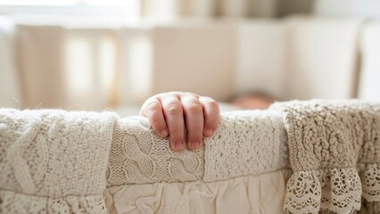 A baby's hand gripping the side of a white crib with textured fabric and a blurred background seen indoors