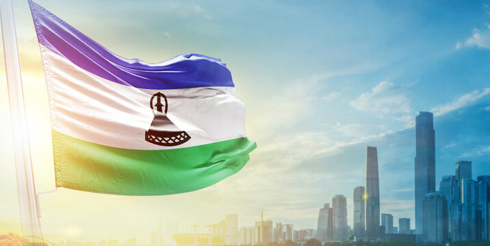 Lesotho flag wavering in beautiful clouds with building skyline. The flag with drone shot in beautiful sky. Lesotho national day flag for independence day. - Powered by Adobe