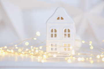 Small white ceramic house with glowing warm fairy lights decoration on soft bokeh background
