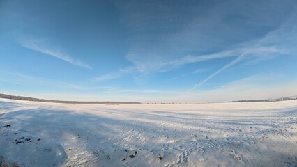 Obraz premium A wide, peaceful winter landscape featuring a snow-covered field under a bright sky. The untouched snow creates a smooth, white surface, surrounded by distant trees and soft natural light.