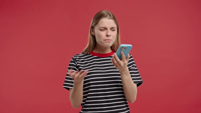Annoyed young pretty woman quarreling while talking on mobile phone. Angry lady in stress. Red studio background. Conflict, dispute, negative