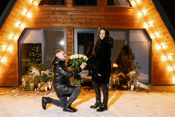 Man kneeling and offering white roses to woman in front of a wooden house at night