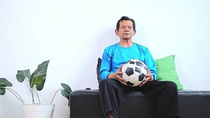 Frustrated older asian man watching soccer game while holding ball at home - Powered by Adobe