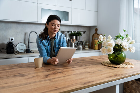 Smiling younger woman using digital tablet in home kitchen, checking online recipes and planning healthy meals while enjoying calm morning routine with coffee before starting her remote freelance work - Powered by Adobe