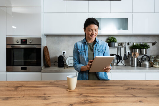 Smiling younger woman using digital tablet in home kitchen, checking online recipes and planning healthy meals while enjoying calm morning routine with coffee before starting her remote freelance work
