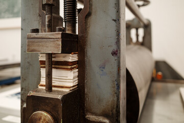 A detailed view of a heavy metal printing press in a workshop setting. The image focuses on the threaded screw and a stack of shims used to adjust the pressure of the roller.