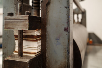 A detailed view of a heavy metal printing press in a workshop setting. The image focuses on the threaded screw and a stack of shims used to adjust the pressure of the roller.