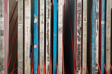 A close-up side view of used screen printing frames stored vertically in a red metal rack within an art studio. The aluminum and wooden frames are stained with dried blue and pink ink. 